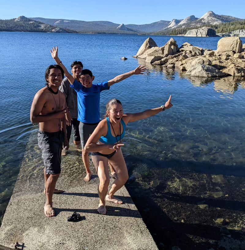 A group of four young people are standing on a concrete platform next to a lake. The water is clear and calm, and there are rocks and trees in the background. The sky is blue and sunny. The people are smiling and appear to be enjoying themselves. One of them is pointing towards the lake.